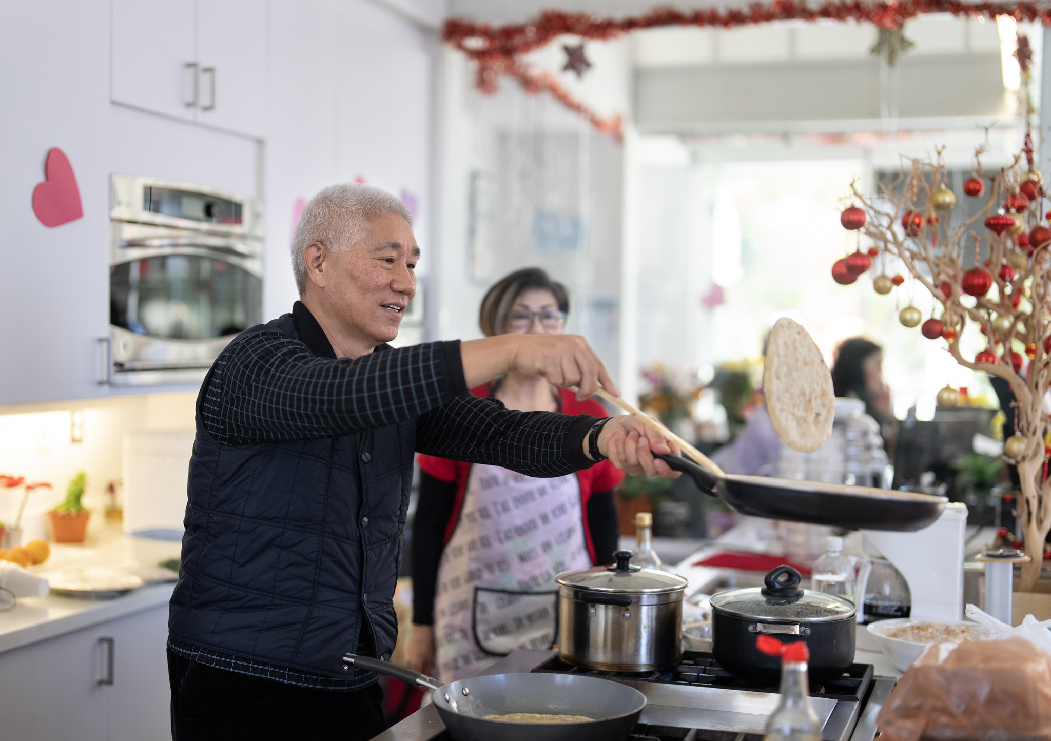 A person cooking in a kitchen with another person standing nearby. Pots and utensils are on the counter.