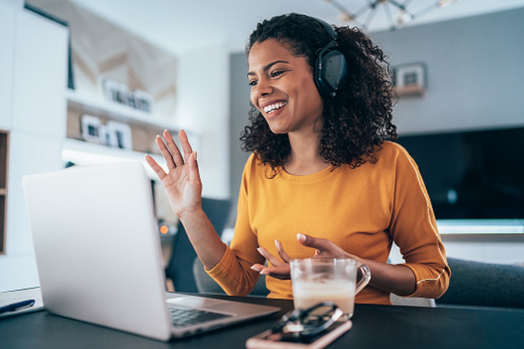 A woman with curly hair smiles and waves during a virtual meeting, promoting community engagement and connection.